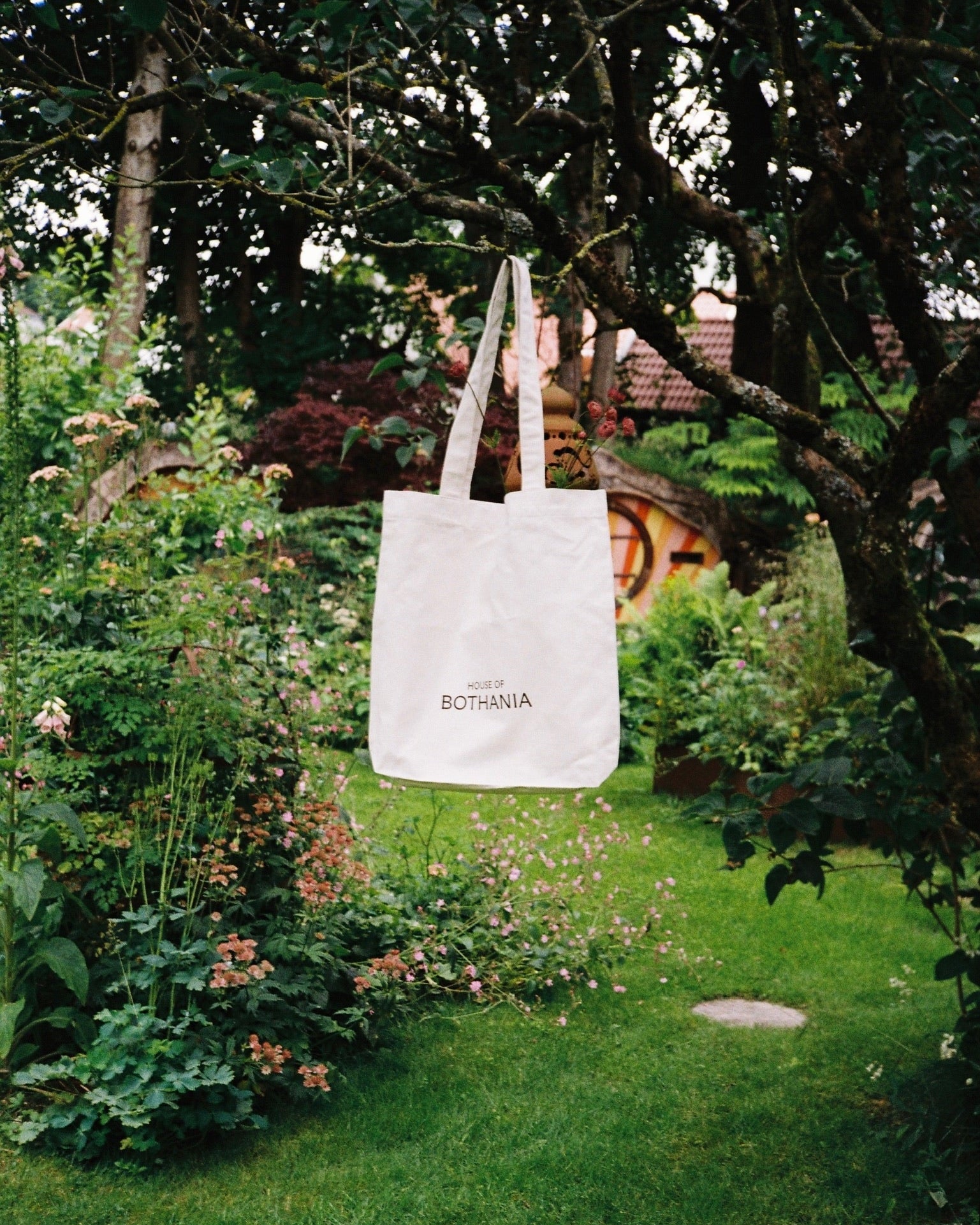 White natural tote bag with House of Bothania' logo, hanging from a tree in a green garden
