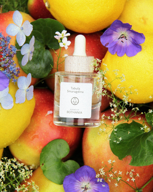 Bottle of the perfume oil 'Tabula Smaragdina' made by House of Bothania. Glass bottle with a wooden cap, on a background of apples and lemons, yuzu with purple and small white flowers. combined with green leaves from lilies as well. Bio-energetic perfume oil made to awaken our senses. Tabula Smaragdina connects us to the throat chakra. Awakens intuition. Evokes clear and peaceful communication. Speak your truth with integ