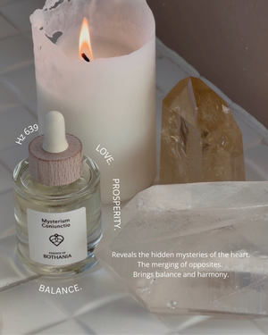 Bottle of Mysterium Coniunctio with a lit candle and crystals on a neutral background. Bio-energetic perfume oil made by House of Bothania, made to awaken your senses. 'Mysterium Coniunctio' is connected to our heart chakra, is represents balance, love and prosperity. It reveals the hidden mysteries of the heart. The merging of opposites. Brings balance and harmony.